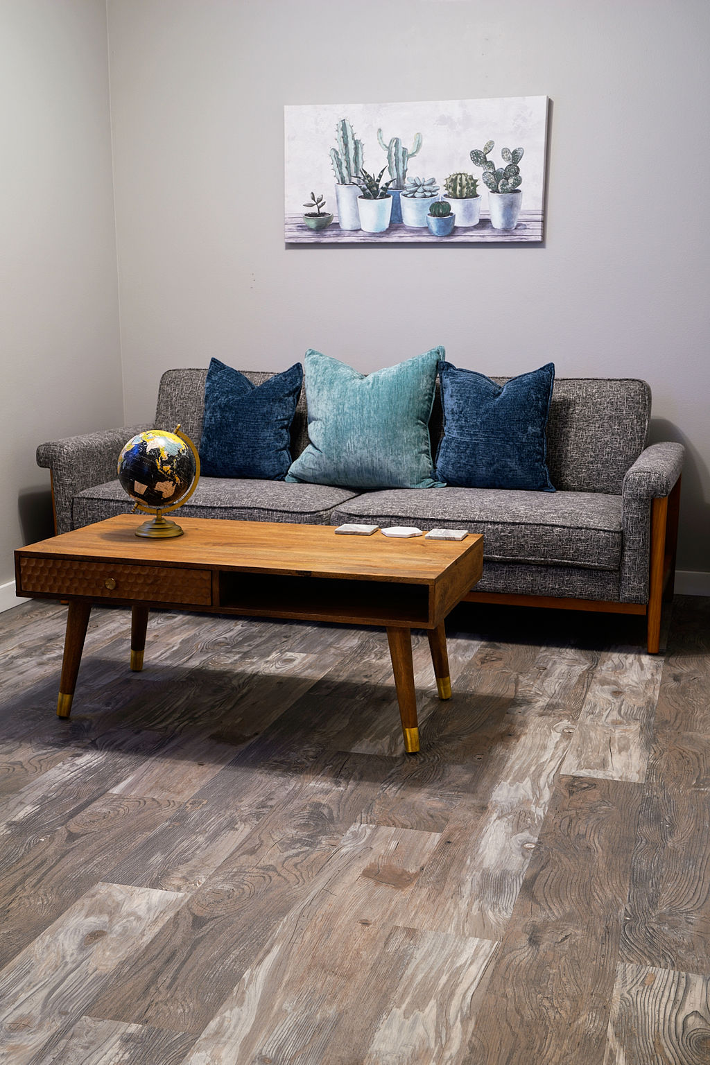 A living area features a gray sofa adorned with teal and navy pillows. A wooden coffee table is positioned in front, accompanied by a decorative globe. Above the sofa, a wall art piece displays a collection of potted plants on a shelf.