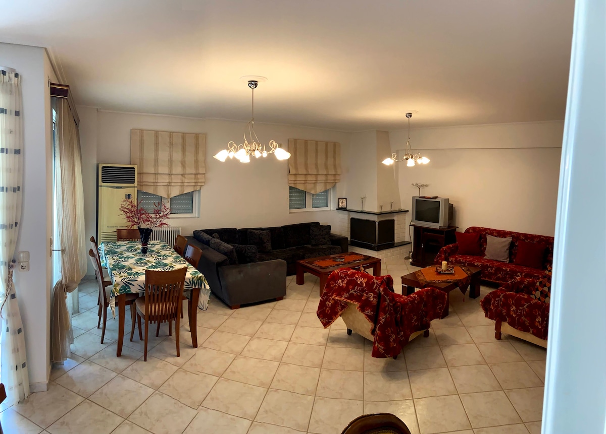 A spacious living area features multiple seating arrangements, including a dark sectional sofa and floral-patterned armchairs. A wooden dining table with matching chairs is situated near large windows. Soft lighting from ceiling fixtures enhances the open layout, while tile flooring provides a clean aesthetic.