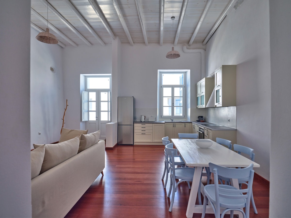 The open-plan kitchen and living area is characterized by high ceilings and wooden floors. A spacious white dining table surrounded by light blue chairs is positioned next to the kitchen, which includes modern appliances. A cozy sofa offers seating, with natural light streaming through the windows.