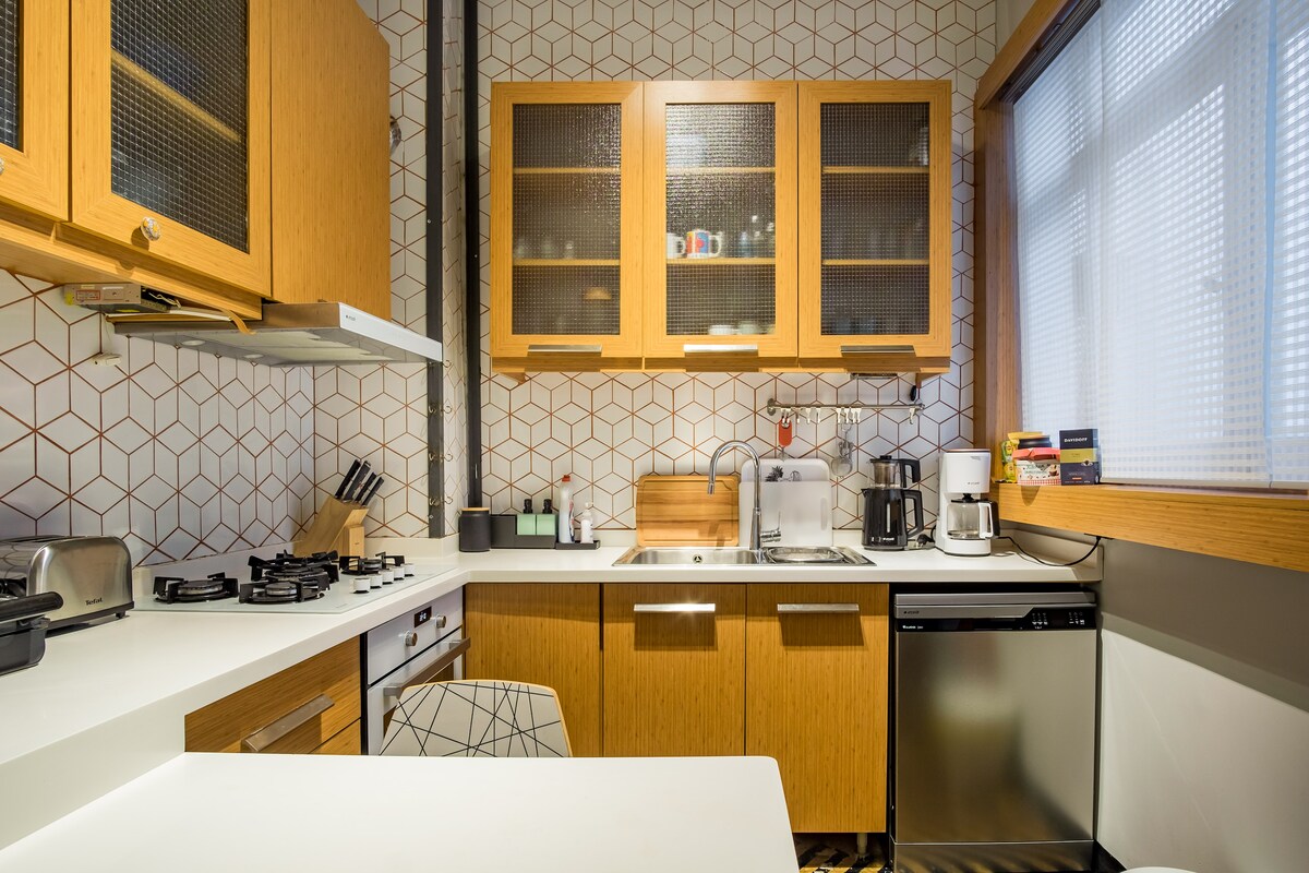 The kitchen features wooden cabinetry with glass panels and modern appliances. A white countertop complements the geometric tile backsplash. Cooking essentials such as a stove, toaster, and coffee maker are neatly arranged, providing convenience for meal preparation in a functional setting.