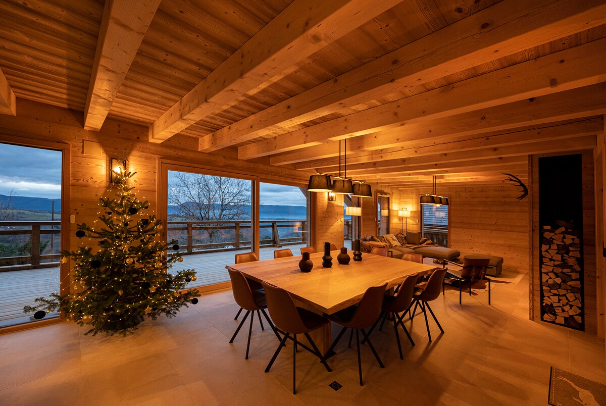 A spacious dining area is illuminated by pendant lights above a large, rectangular wooden table surrounded by several chairs. A decorated tree stands in the corner, with large windows offering views of the surrounding landscape. Wooden beams and natural materials are present throughout the room.
