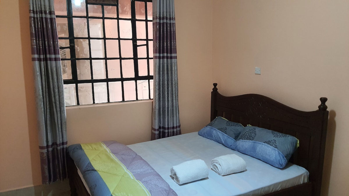 The room features a spacious bed adorned with colorful blankets and neatly arranged towels. Large windows allow natural light to fill the space, framed by light-colored curtains that add to the room's brightness.