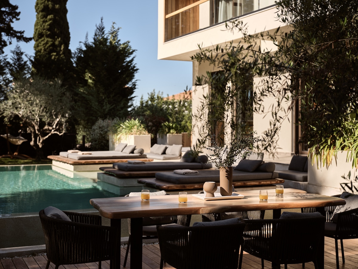 An outdoor dining area features a wooden table surrounded by black chairs, set against a backdrop of a private pool with sunbeds. Soft green landscaping complements the scene, while candles and a decorative vase enhance the inviting atmosphere.