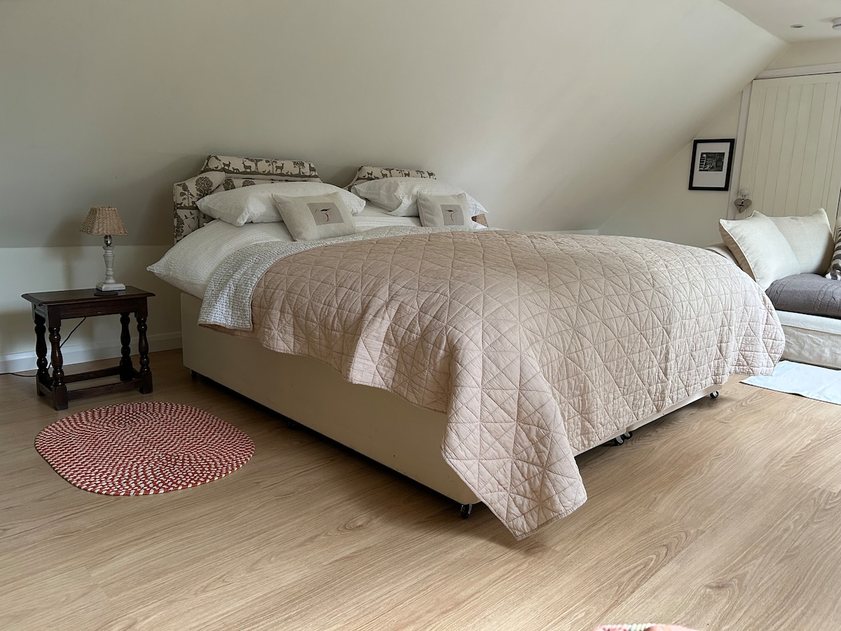 A spacious bed is positioned against a sloped ceiling, covered with a quilted beige bedspread. Pillows are neatly arranged, and a small bedside table with a lamp is situated to one side. A cozy seating area with a light-colored sofa is visible in the background.