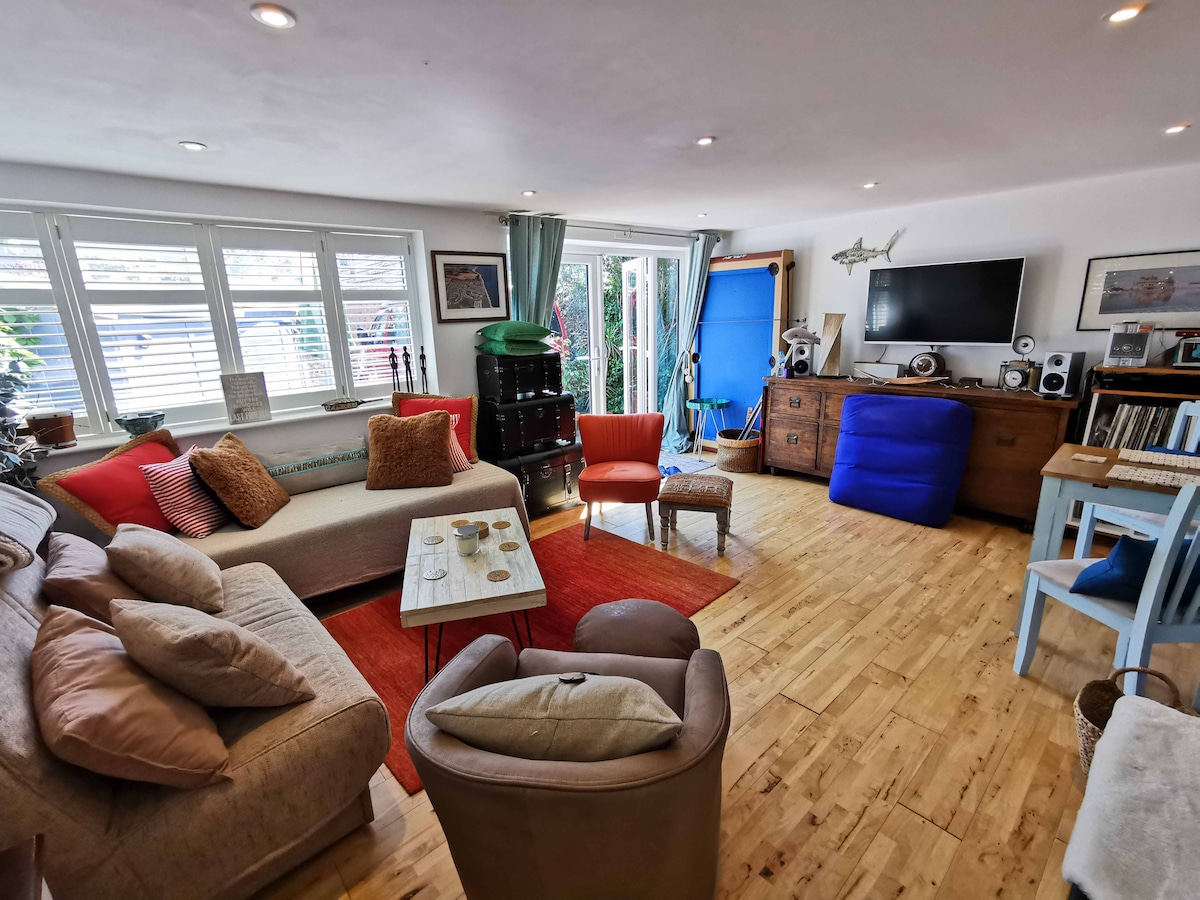 A welcoming living area features a mix of seating options, including a cozy sofa adorned with cushions, a colorful armchair, and a bean bag. Natural light fills the space through multiple windows, while a wooden table is set with decorative items. A media unit stands opposite the seating.