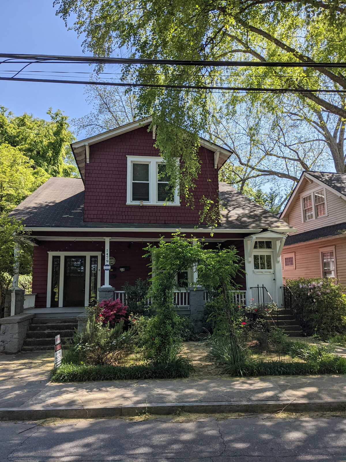 A charming triplex house is shown, featuring a maroon exterior with white trim. Lush greenery surrounds the property, including flowering plants and trees. A porch with steps leads to the main entrance, complemented by well-maintained landscaping along the pathway.