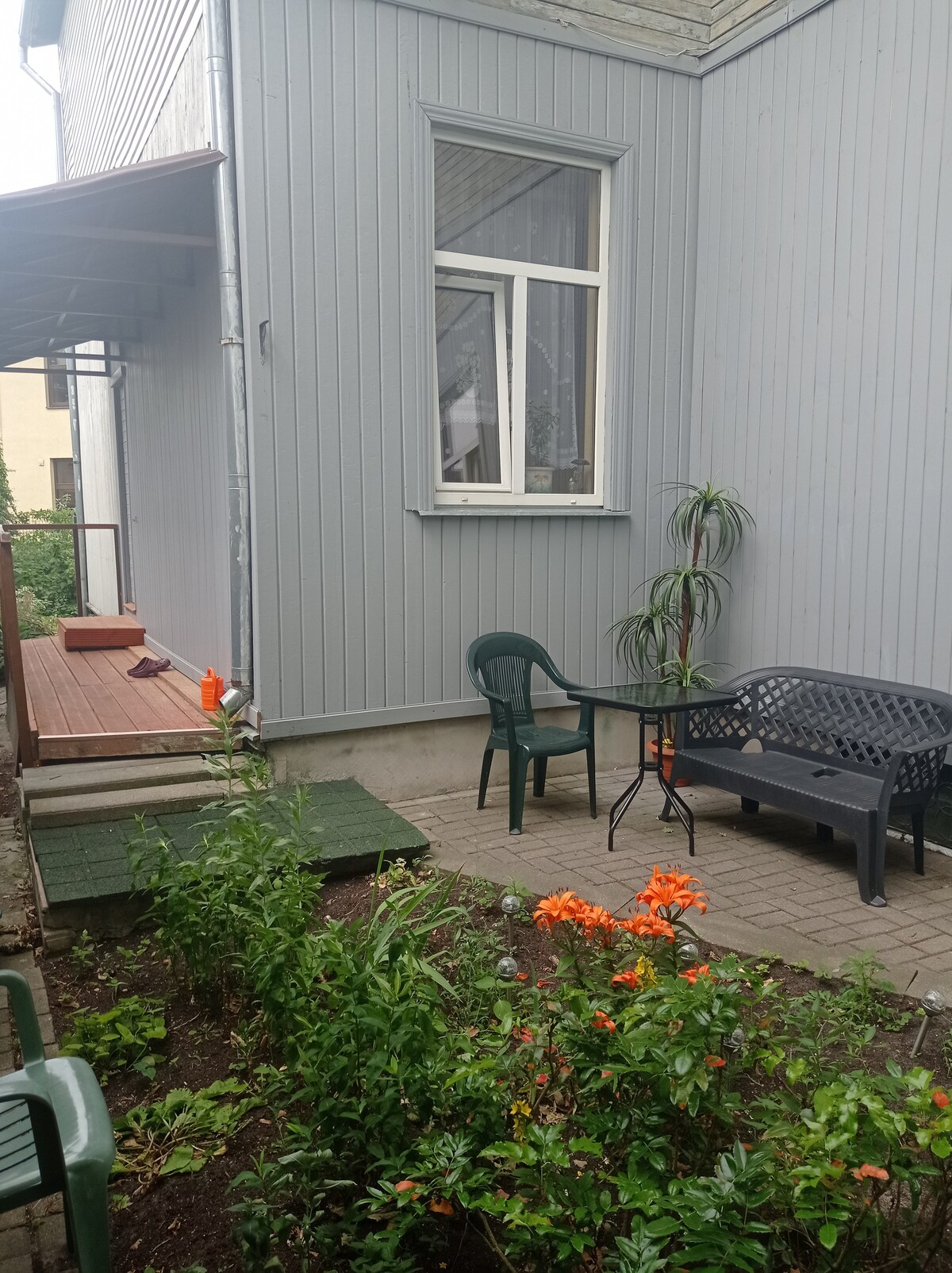 An outdoor seating area is set in a small garden, featuring a table and chairs. Bright orange flowers contrast with the greenery. Gray wooden siding is visible on the building, alongside a window that allows natural light into the space.