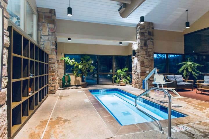 An indoor pool area is framed by large windows that provide natural light. Stone pillars support the ceiling, while potted plants add greenery. A set of loungers is arranged nearby, and the pool features steps leading into the water, inviting relaxation.