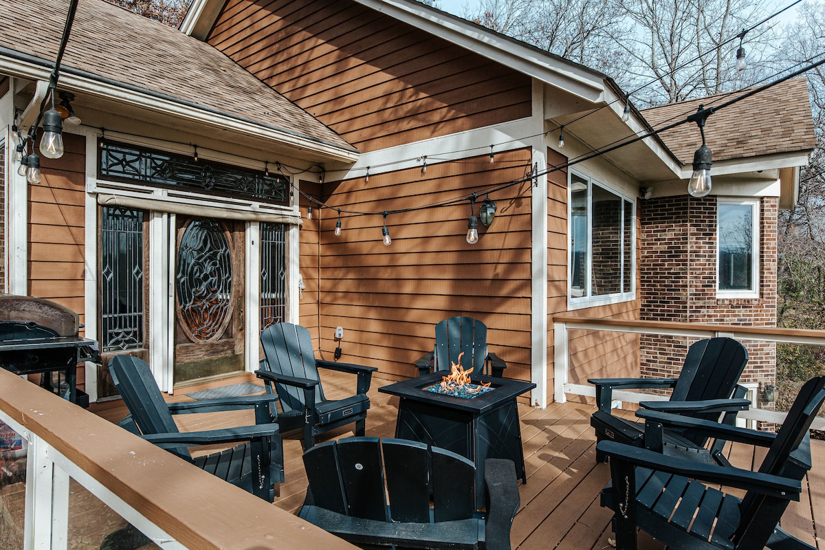 A spacious deck area features several Adirondack chairs arranged around a central firepit. Dimmable string lights enhance the ambiance. A gas barbecue grill is positioned nearby, and a large decorative door with intricate glasswork adds character to the space.