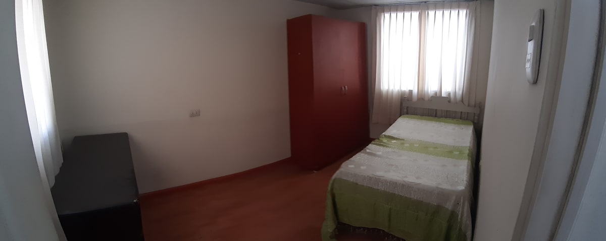 A minimalist bedroom is presented, featuring a spacious bed with a light green spread. Natural light floods through the vertical blinds of a window. A medium-sized wardrobe in a warm hue stands against one wall, with a simple black dresser positioned nearby.