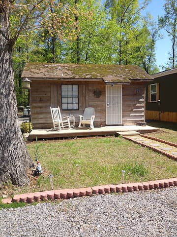 A rustic cabin is framed by greenery, showcasing a porch with two rocking chairs positioned in front of the entrance. The cabin's weathered wooden exterior features a single window adorned with a decorative accent, alongside a well-kept flowerbed bordered by bricks.