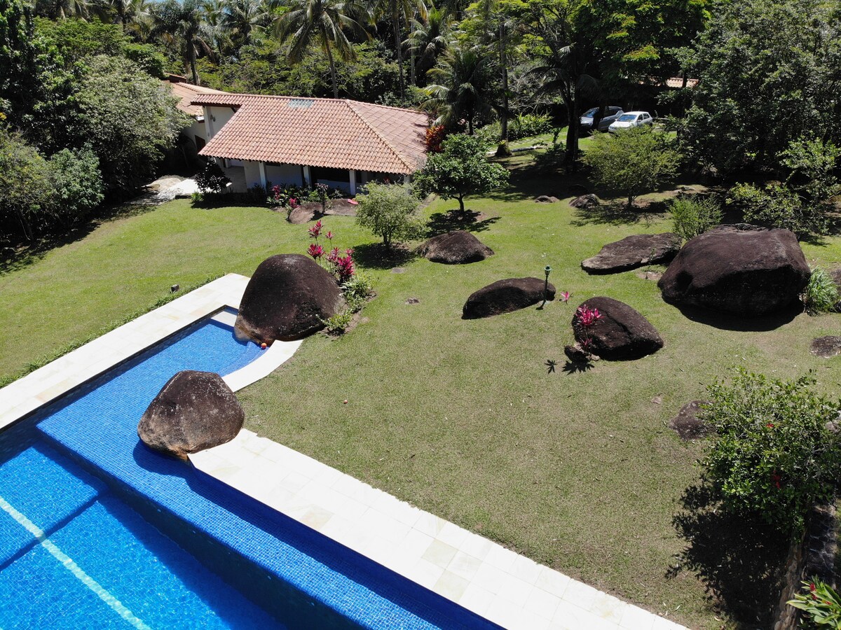 An aerial view of a sprawling property features a large blue swimming pool adjacent to a well-maintained lawn. Natural stones are interspersed throughout the garden, surrounded by lush greenery and flowering plants. A house with a terracotta roof is visible in the background.