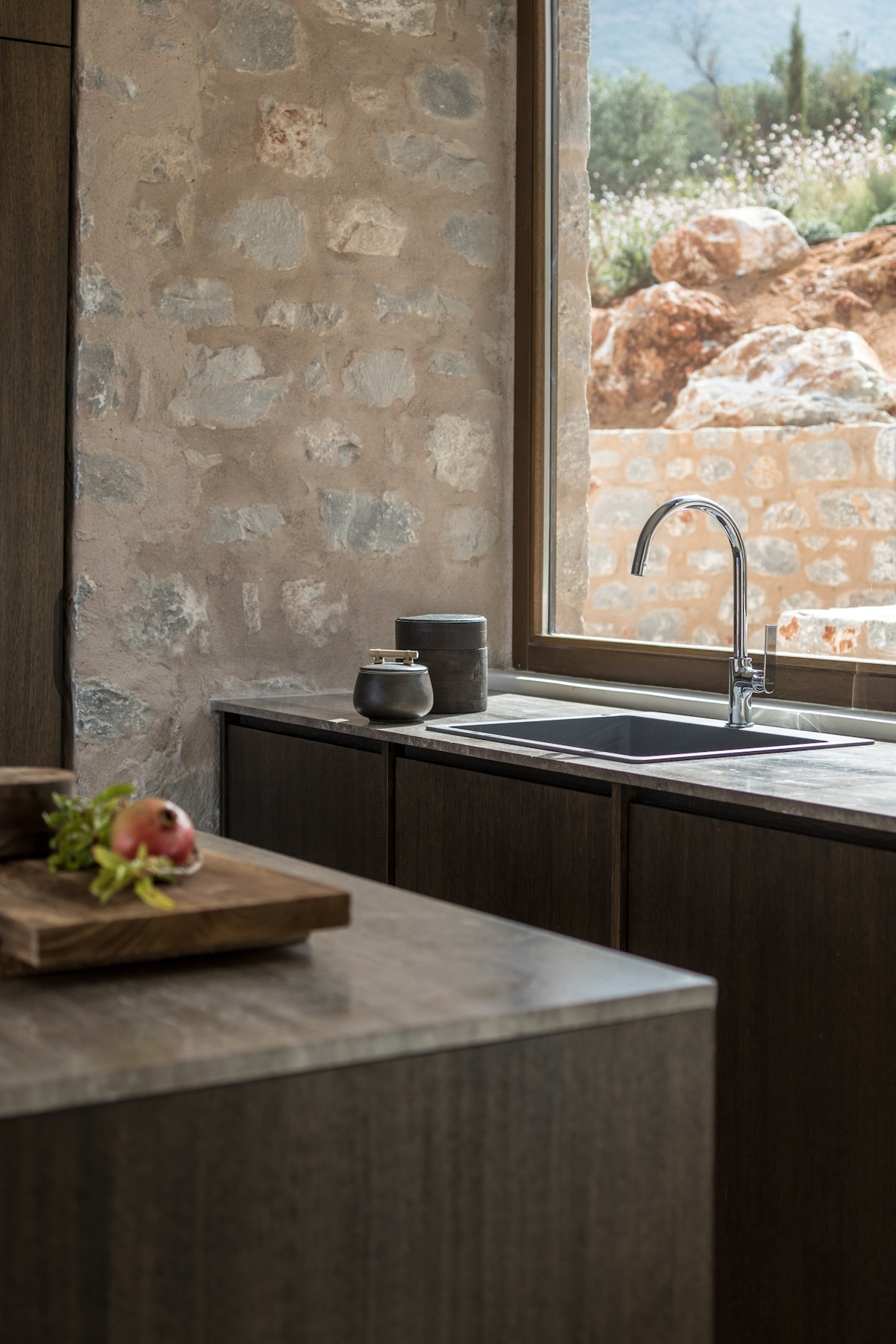 The kitchen features natural stone walls and wooden cabinetry. A large window offers views of the outdoors, while a sleek sink and countertop draw attention. Fresh herbs and a fruit bowl sit on the island, enhancing the space's functional design.