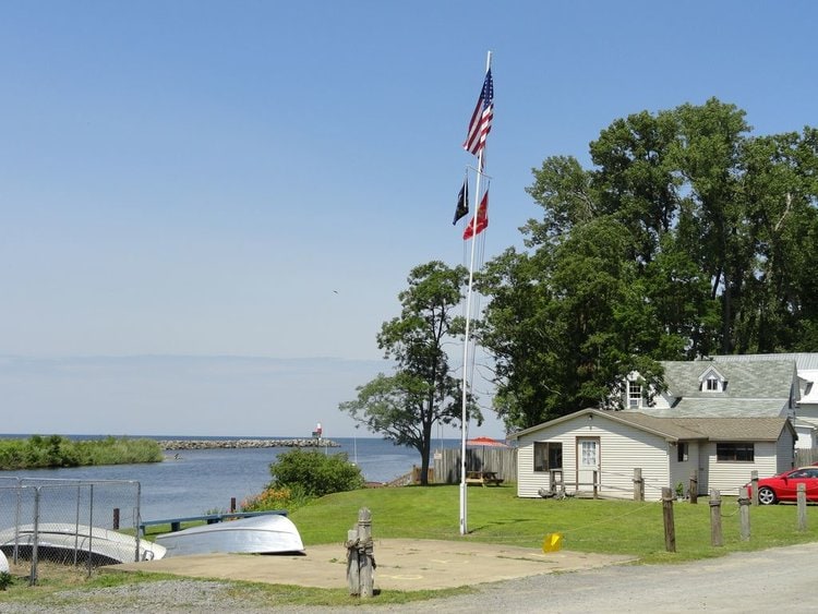 A peaceful waterfront scene captures a grassy area adjacent to the water. A flagpole displays several flags, while a few boats are visible on the shore. The surrounding trees provide a natural backdrop, complementing the serene atmosphere.