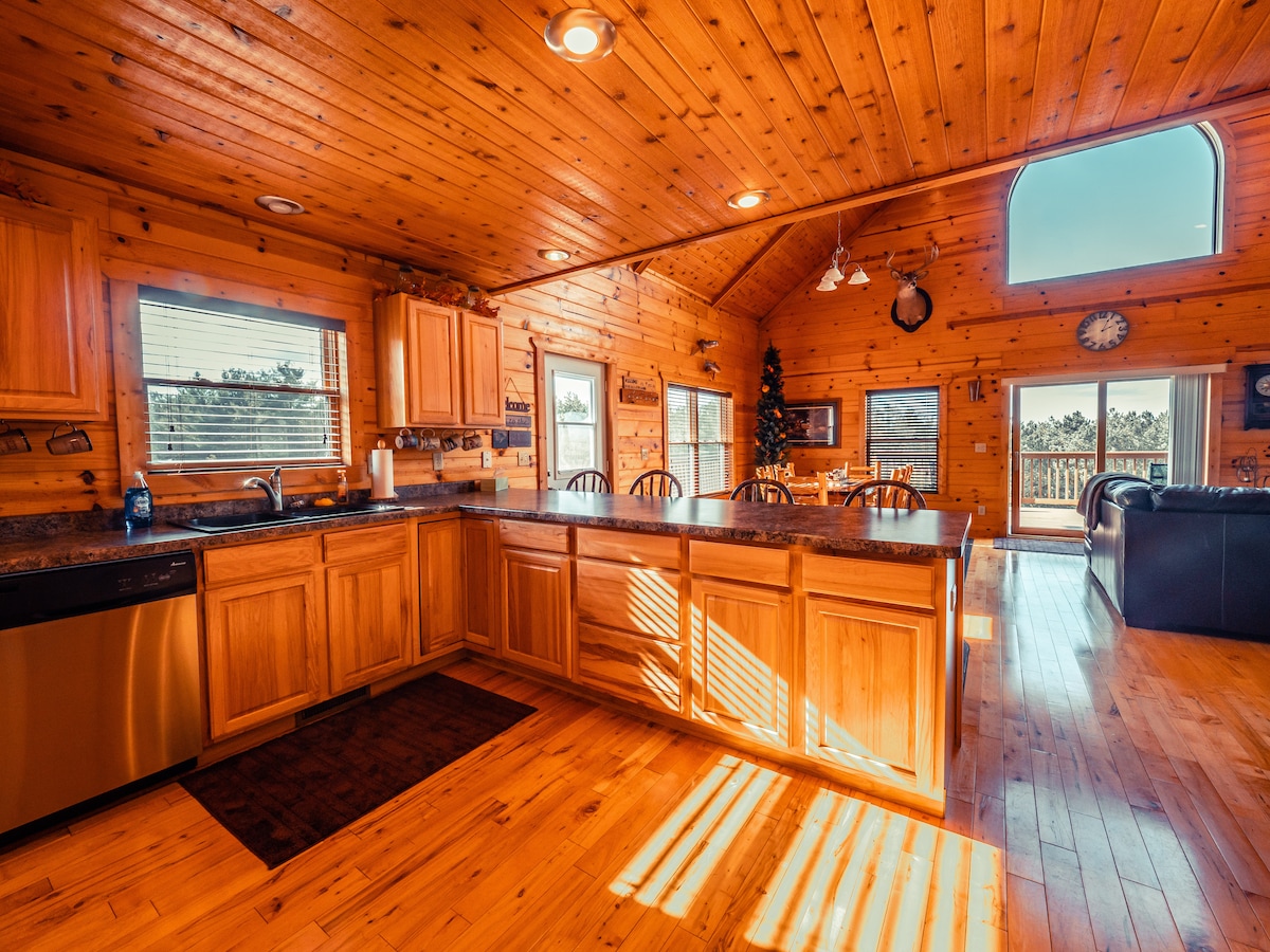 A spacious kitchen is highlighted by wooden cabinetry and a dark countertop. Natural light fills the room through multiple windows, creating a warm ambiance. Open to the living area, this welcoming space features a dining option with several chairs and offers views of the exterior through the nearby door.