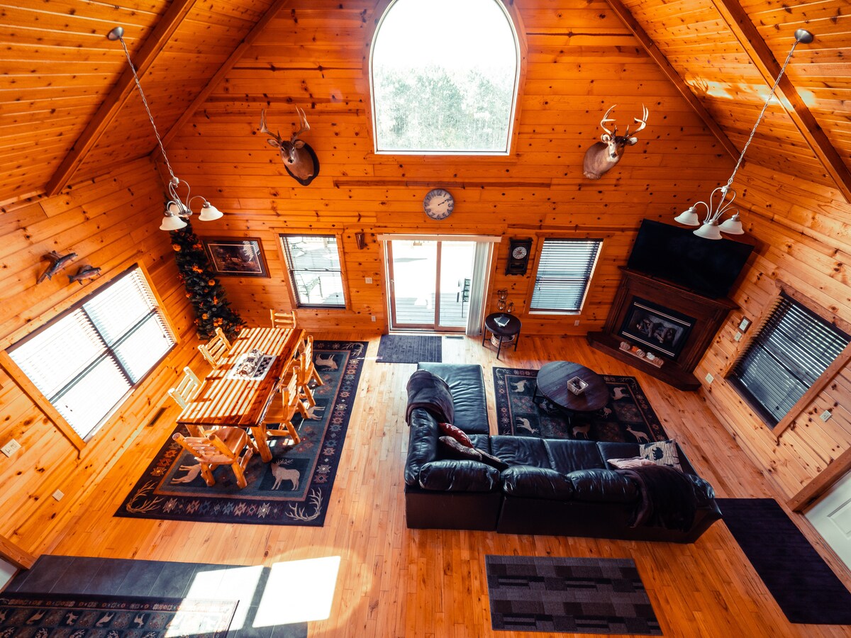 An open living space showcases a high wooden ceiling and warm wood paneling. A dining table with wooden chairs occupies one area, while a black leather sofa and a circular coffee table are positioned around a cozy rug, enhancing the cabin's inviting atmosphere.