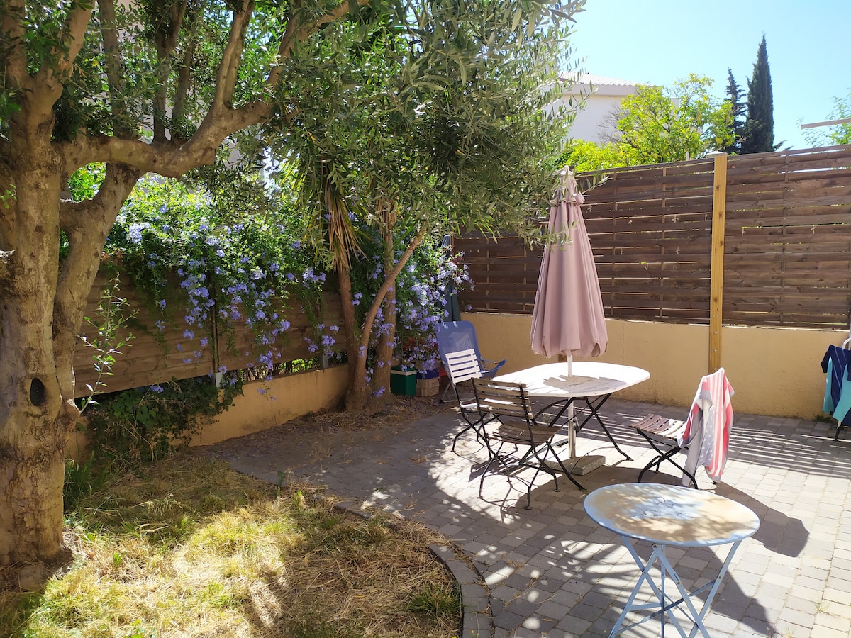 A peaceful garden space features a stone patio surrounded by greenery and colorful flowers. A table with chairs is positioned under a large umbrella, while a blue table adds a pop of color. The area is shaded by an olive tree, providing a serene atmosphere.