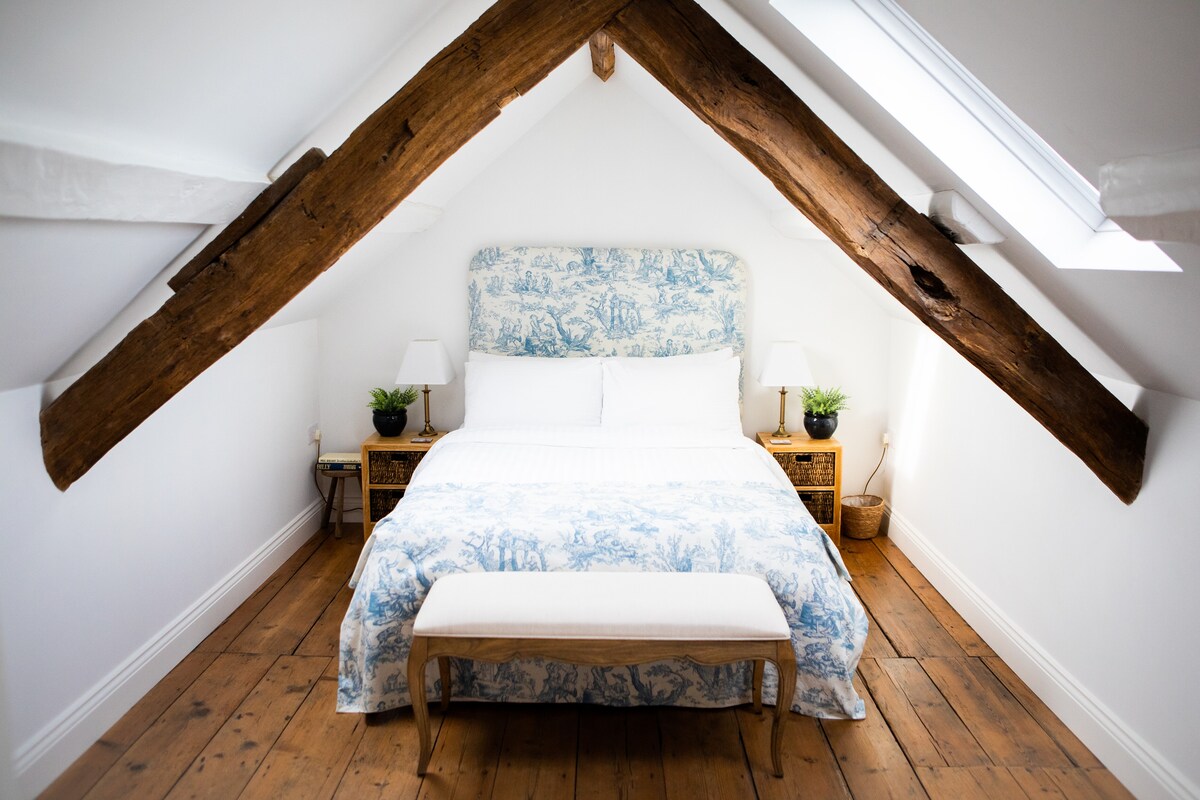 A bright and airy bedroom features a soft blue and white patterned headboard above a spacious bed. Warm wooden beams span the ceiling, complemented by natural light from two skylights. Nightstands with lamps sit on either side of the bed, enhancing the serene atmosphere.