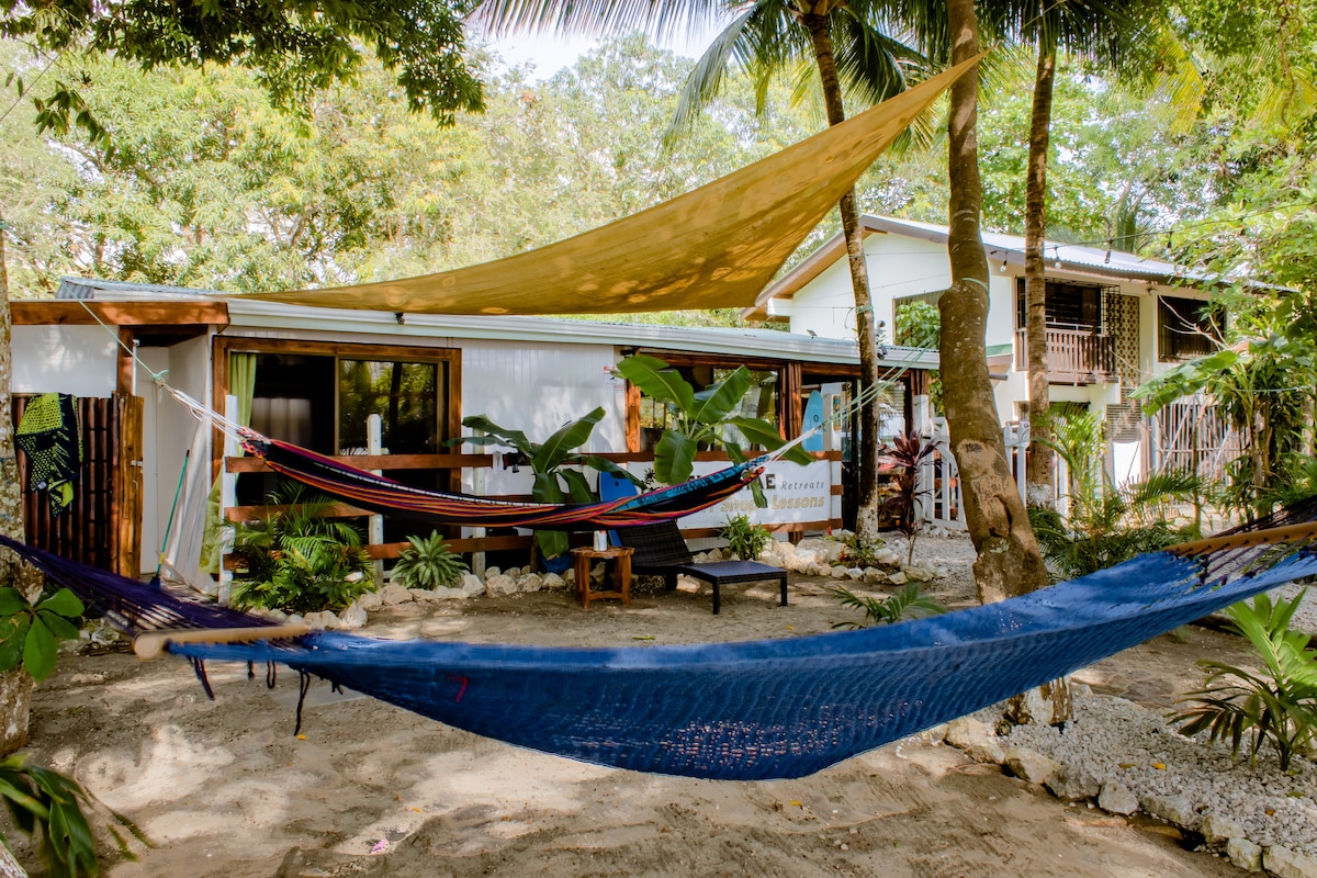 A relaxing outdoor space is showcased with several colorful hammocks suspended between trees. The sandy area is surrounded by lush greenery, creating a serene environment. A small seating area and nearby structures provide a cozy backdrop, inviting guests to unwind in nature.