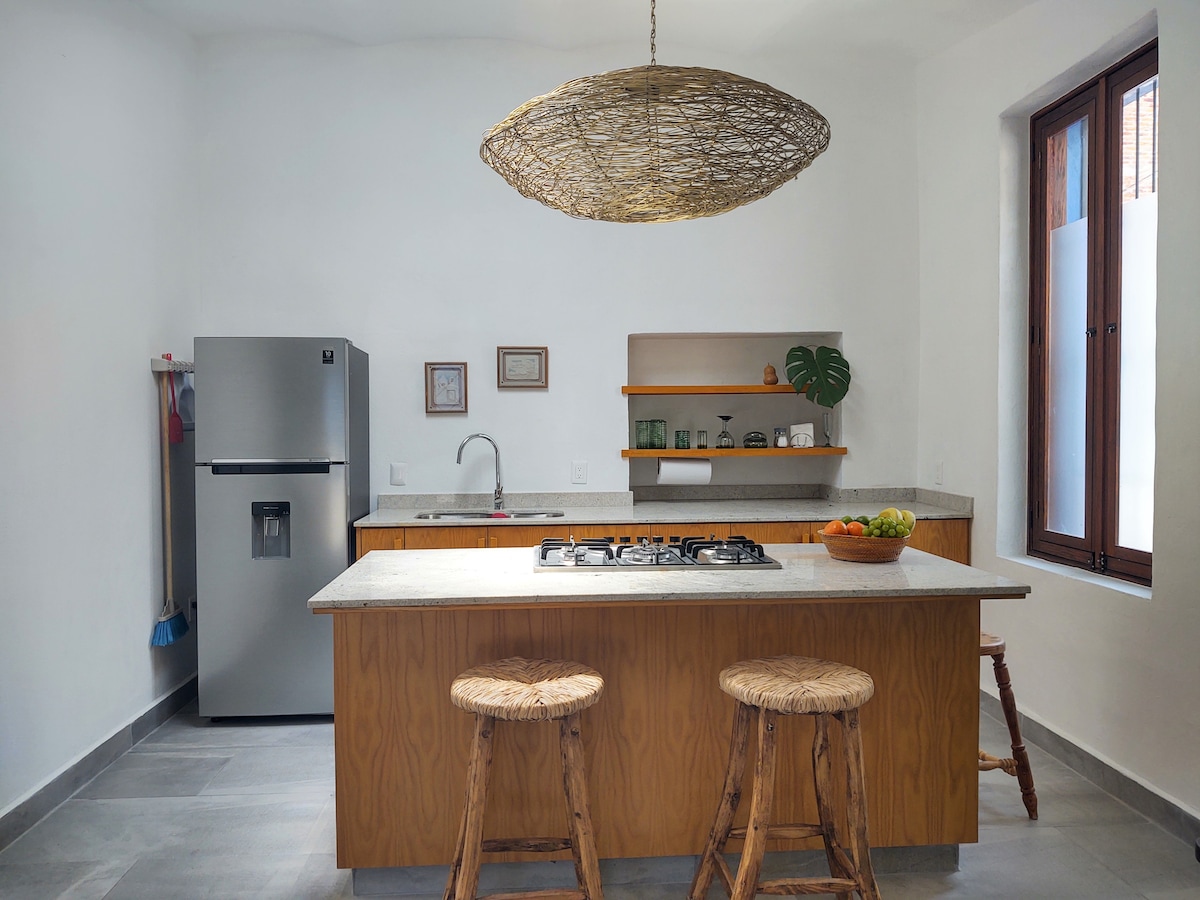 A well-equipped kitchen features a central island with seating for two on natural wooden stools. Light wooden shelves display decorative items, while a stainless steel refrigerator stands adjacent to a sleek countertop. Natural light enters through two windows, highlighting the spacious design.