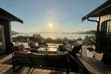 A spacious outdoor seating area is presented, featuring comfortable lounge furniture arranged around a central table. The setting overlooks a serene water view, with soft sunlight illuminating the scene, enhancing the peaceful atmosphere. Potted plants are visible, contributing to the inviting environment.