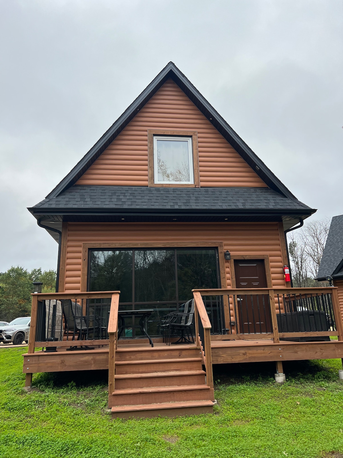 A cabin featuring a traditional A-frame design is shown from the front. Large windows allow natural light to fill the interior, and a wooden deck with multiple chairs is located outside. Steps lead up to the entrance, set against a backdrop of greenery.