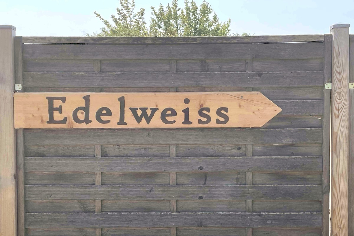 A wooden sign displaying the word 'Edelweiss' is mounted on a fence. The sign features a clear arrow pointing to the right, with natural wood tones contrasting against the weathered grey wood of the fence.