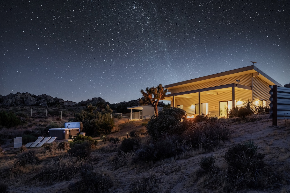 The exterior of the house is showcased under a star-filled sky, highlighting expansive glass doors that open to a patio area. A hot tub is positioned nearby, surrounded by desert foliage and rocks, fostering a serene ambiance.