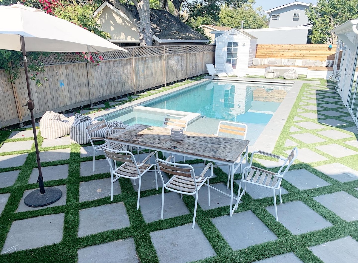 An outdoor area features a swimming pool surrounded by a manicured lawn. A table with several chairs is positioned beside the pool, and lounge chairs are placed nearby. Shade is provided by a large umbrella, enhancing the relaxing outdoor space.