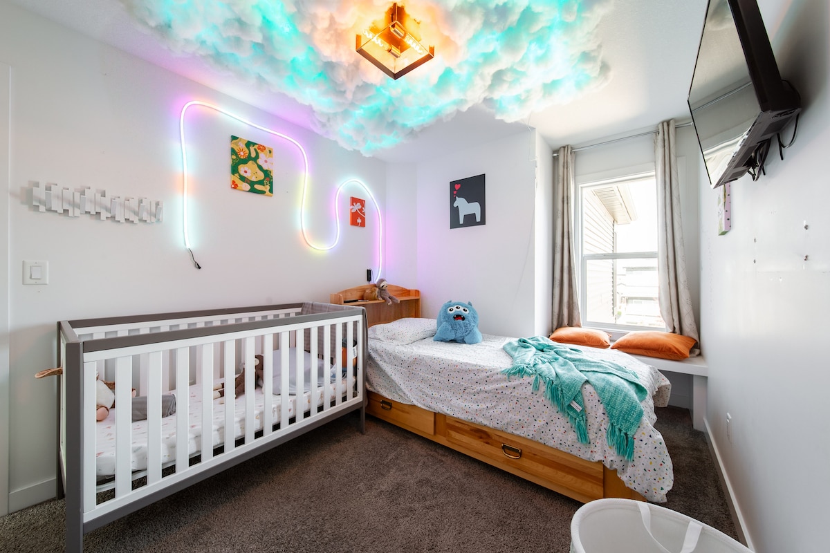 The children's room features a crib and a spacious bed, surrounded by playful decor including cloud-like lighting and vibrant wall art. Natural light fills the room through a window, with a colorful blanket draped over the bed and extra seating on a small nook.