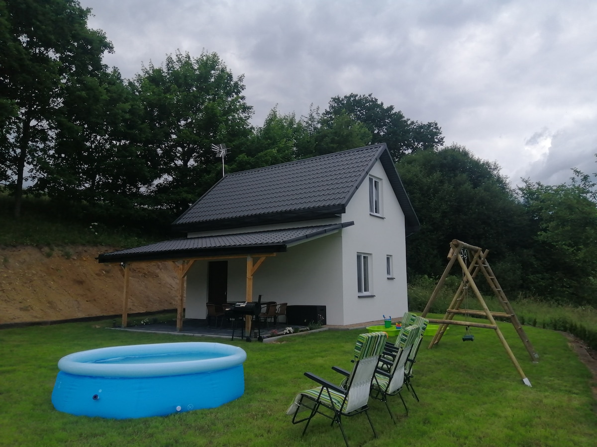 A charming house is positioned amid greenery, featuring a covered porch. An inflatable pool occupies the grassy area, while a swing set and several lounge chairs are nearby, providing inviting spaces for relaxation and play.