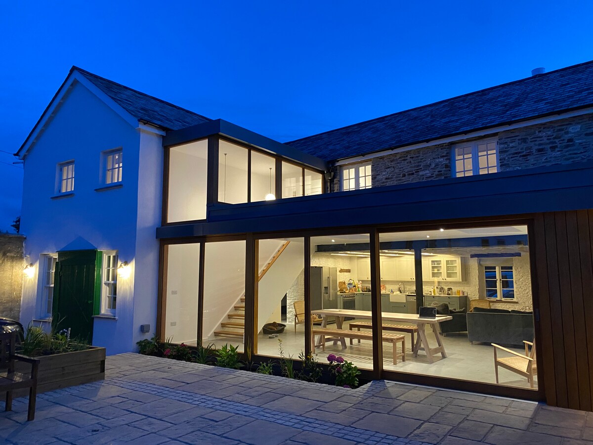 A charming exterior view of the renovated sail loft is presented at dusk, featuring large glass doors that open to an inviting interior space. The building showcases a mix of stone and modern finishes, with warm lights visible through the windows and a vibrant door on the ground floor.