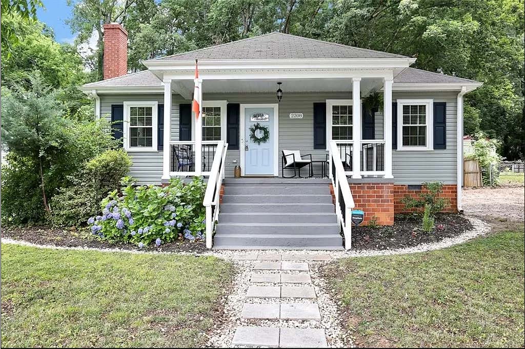 A newly renovated home is presented with a wide front porch featuring two black chairs. Lush green foliage and colorful flowers adorn the landscape, enhancing the entrance. A pathway of stones leads to the front door, framed by blue shutters on the windows.