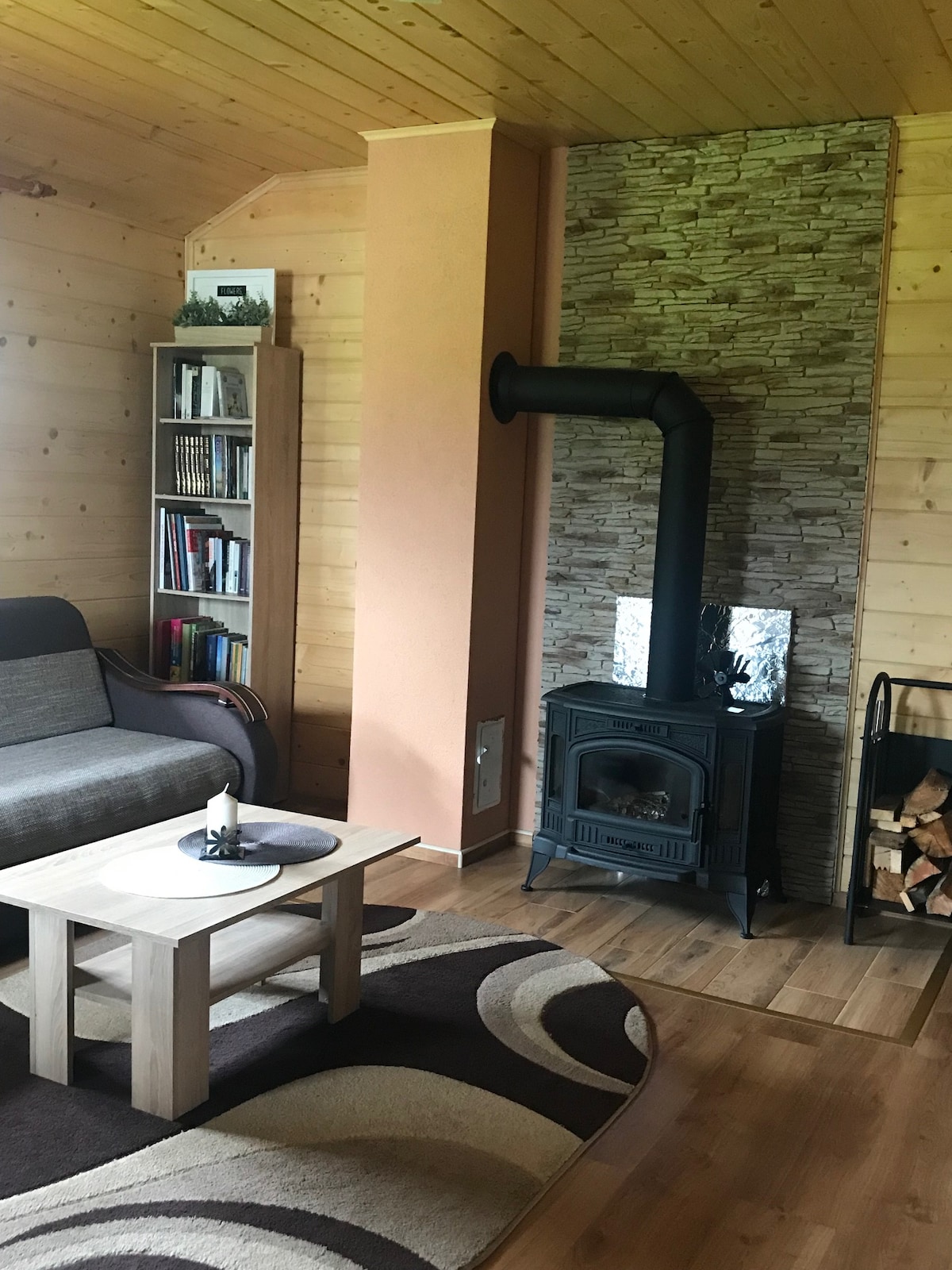 A cozy living area features a gray sofa paired with a wooden coffee table. A modern wood stove is positioned against a textured stone wall, complemented by a neatly stacked pile of firewood. A rug with a circular pattern adds warmth to the wooden floor.
