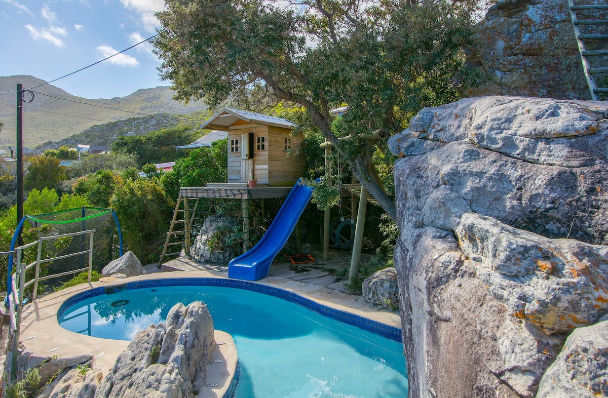 A swimming pool is surrounded by natural rock formations, with a wooden playhouse nestled in the trees nearby. A blue slide extends from the playhouse, offering fun for children. The area is shaded by lush greenery, with a swing set visible in the background.