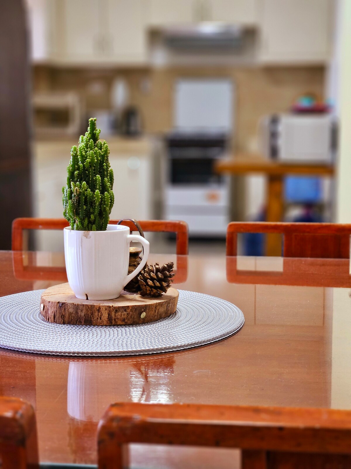 A dining table set is visible with wooden chairs surrounding it. On the table, a white cup and a small plant are positioned on a wooden slice, complemented by a pinecone. The kitchen area can be seen in the background.