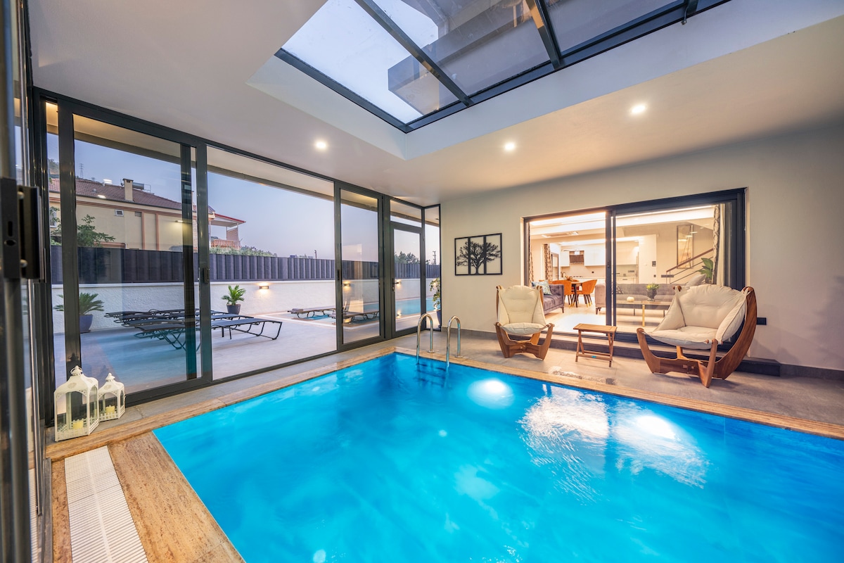 An indoor pool is featured, surrounded by floor-to-ceiling glass doors that provide a view of the outdoor area. Two comfortable chairs are placed nearby, with natural light streaming through the skylight above, creating a bright and inviting space.