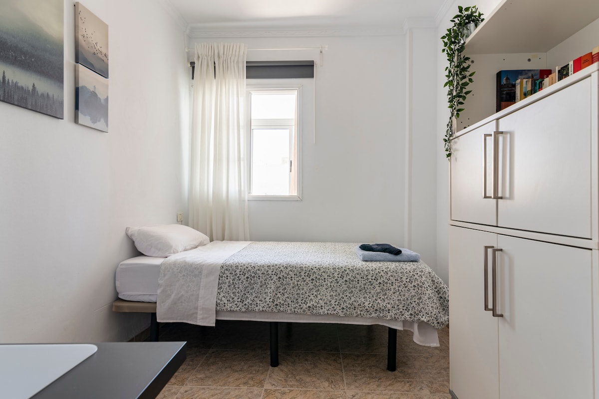 A private bedroom is filled with natural light, featuring a single bed dressed with a light patterned coverlet. A tall shelf is visible, offering organized books and a decorative plant. The window is dressed with sheer white curtains, maintaining a bright and airy feel.