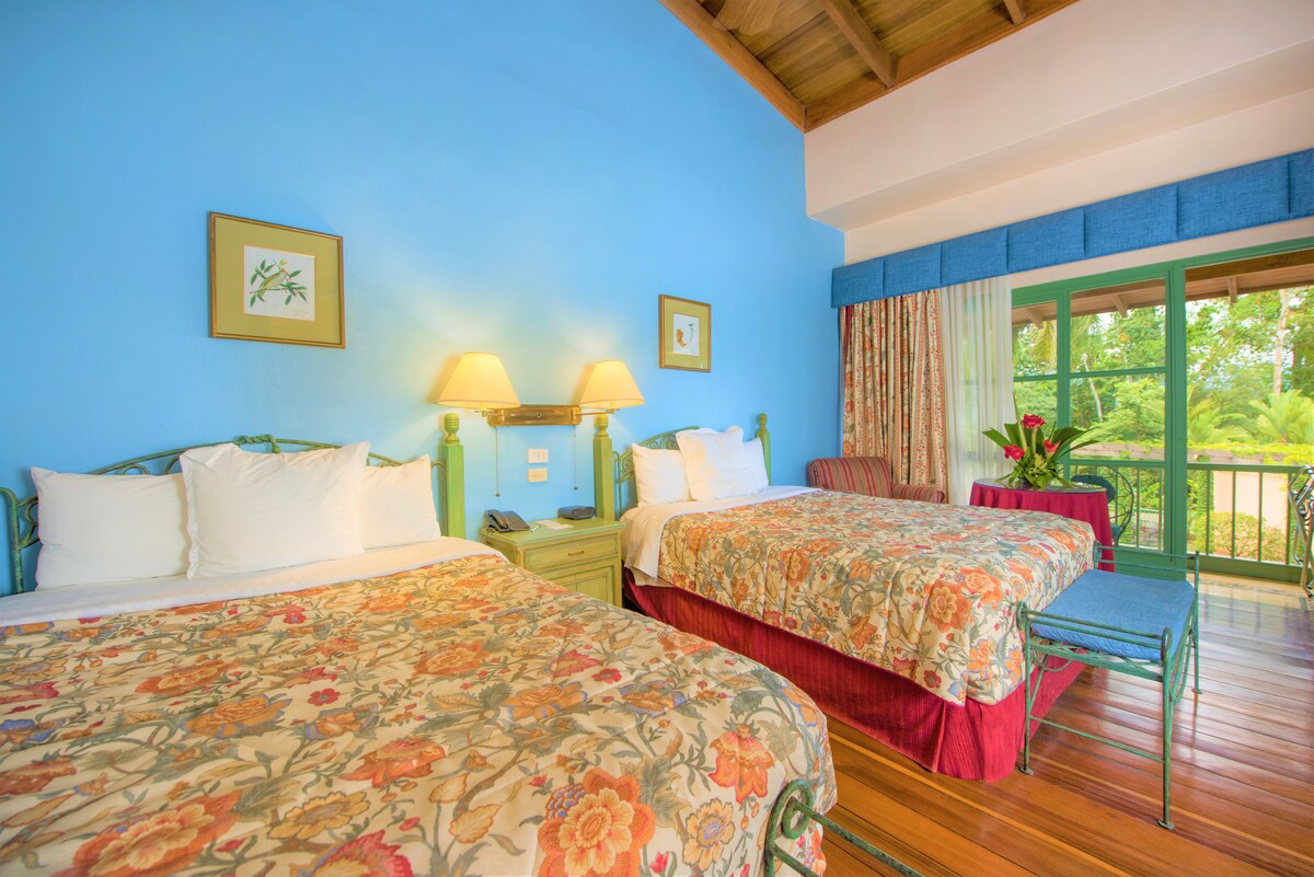 The room features two queen beds adorned with floral bedspreads and light-colored pillows. A small table and chairs are positioned near the window, which offers views of the garden. The walls are painted in soothing blue, complemented by natural wood finishes.