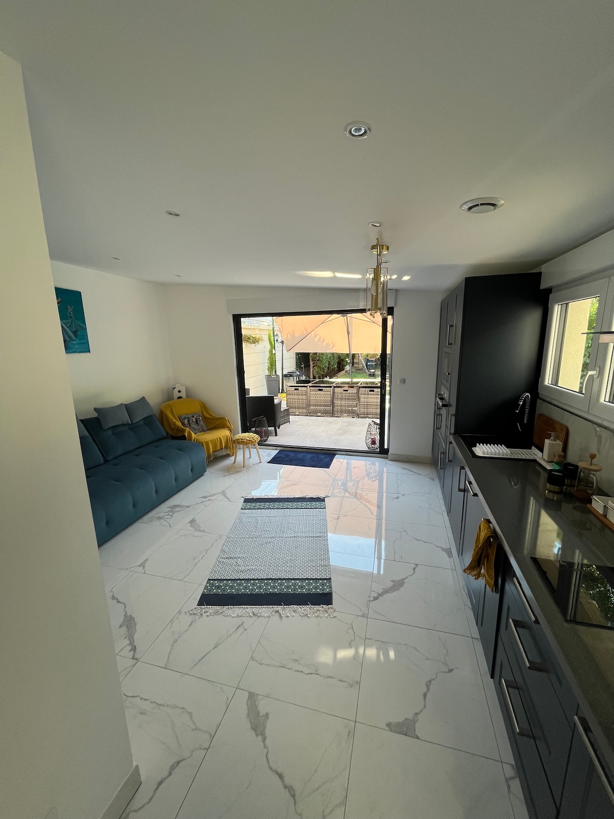 A bright and spacious studio is displayed, featuring a comfortable sofa, minimalist decor, and large windows that open to a terrace. Light-colored cabinetry complements the marble flooring, while a decorative rug adds warmth. Outdoor seating is visible through the glass doors.