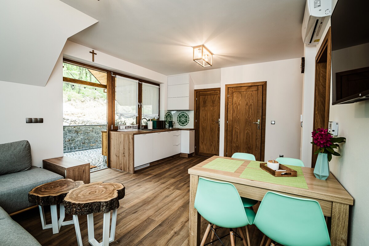 An open living area features a light wood dining table surrounded by aqua-colored chairs. A gray sofa is positioned nearby. The kitchen showcases modern appliances and a unique circular window. Natural light floods the space through large glass doors leading to an outdoor area.