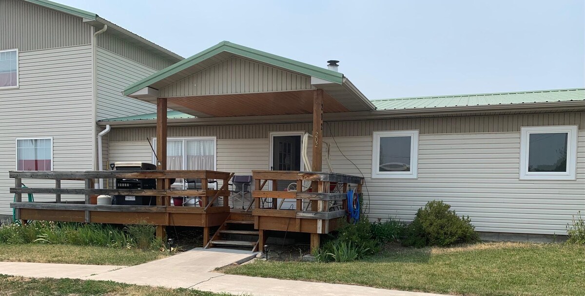 A private entrance leads to a wooden deck, featuring outdoor seating and a barbecue grill. The home is positioned beside a well-maintained lawn, with a pathway leading up. Large windows are visible on the side, allowing natural light into the interior.