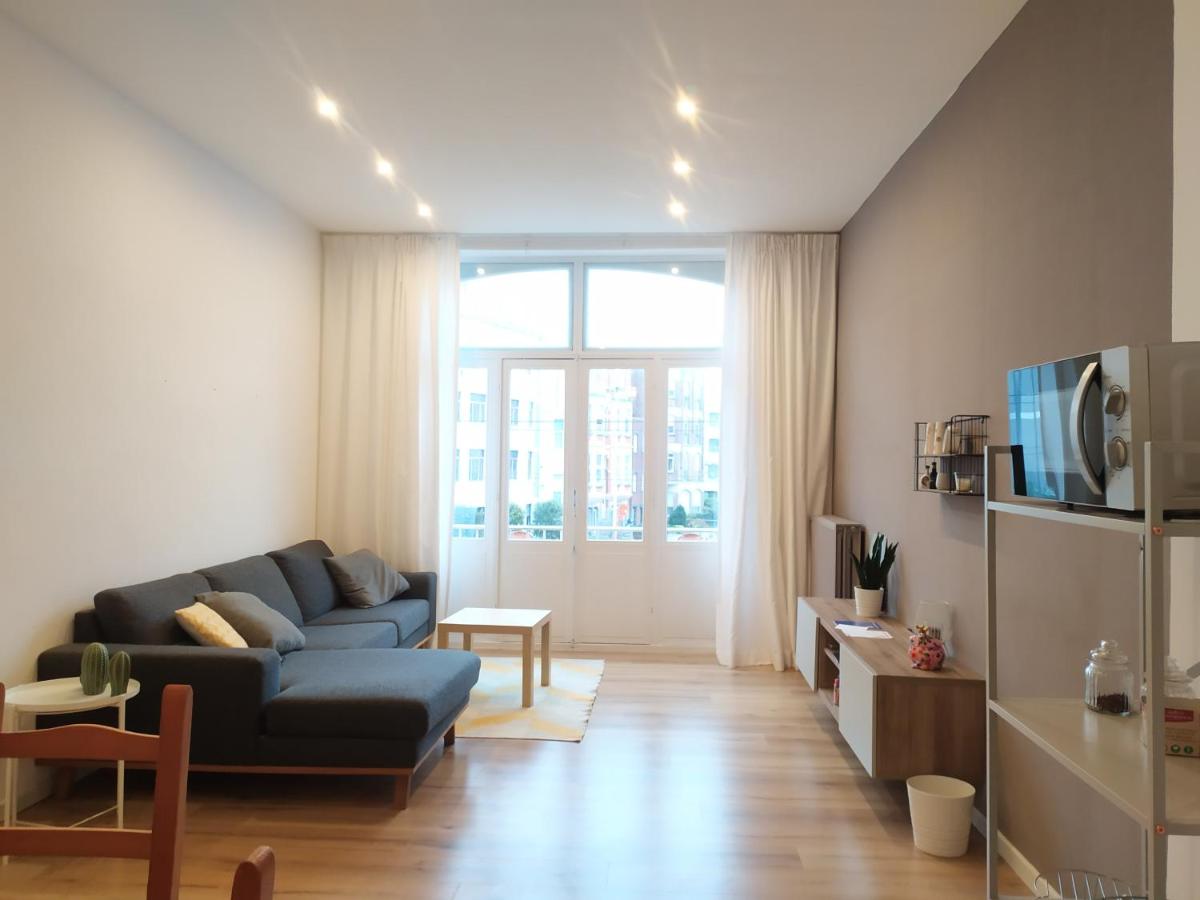A light-filled living area is presented with a comfortable gray sofa and a wooden coffee table. Large windows allow natural light to brighten the space. A simple shelving unit holds decorative items, while a glimpse of greenery is visible outside. The room features warm wooden flooring.