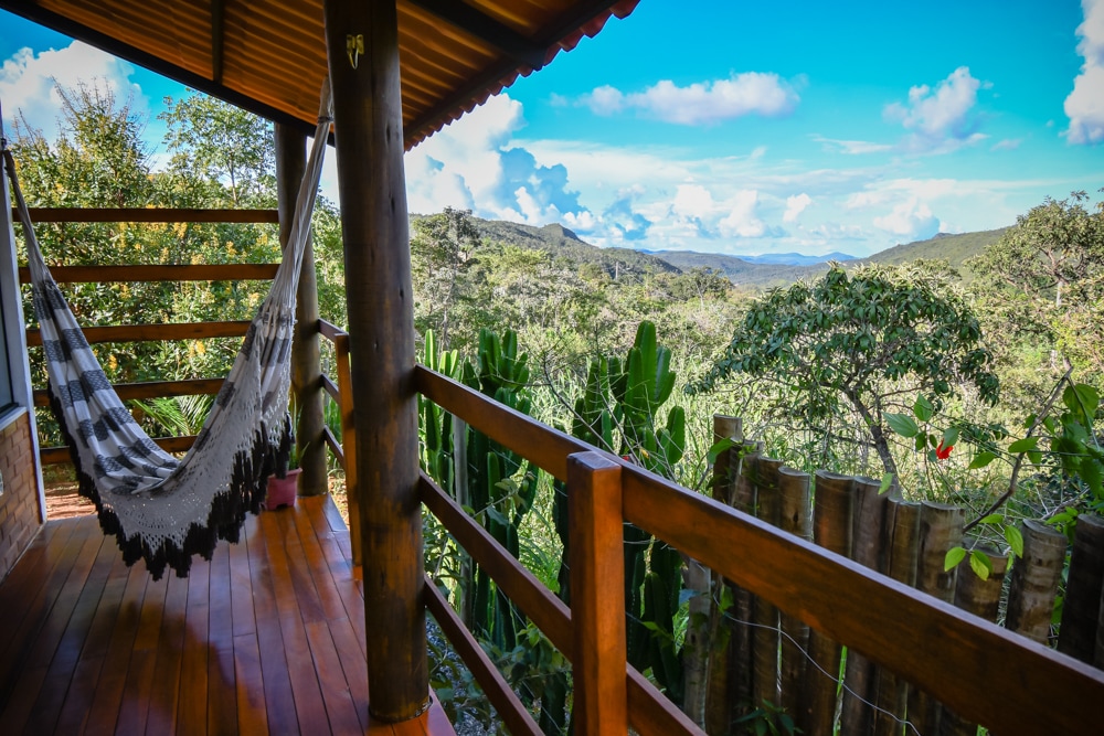 A wooden deck features a traditional hammock, providing a comfortable spot for relaxation. Lush greenery and rolling hills extend into the distance, under a bright blue sky adorned with scattered clouds.