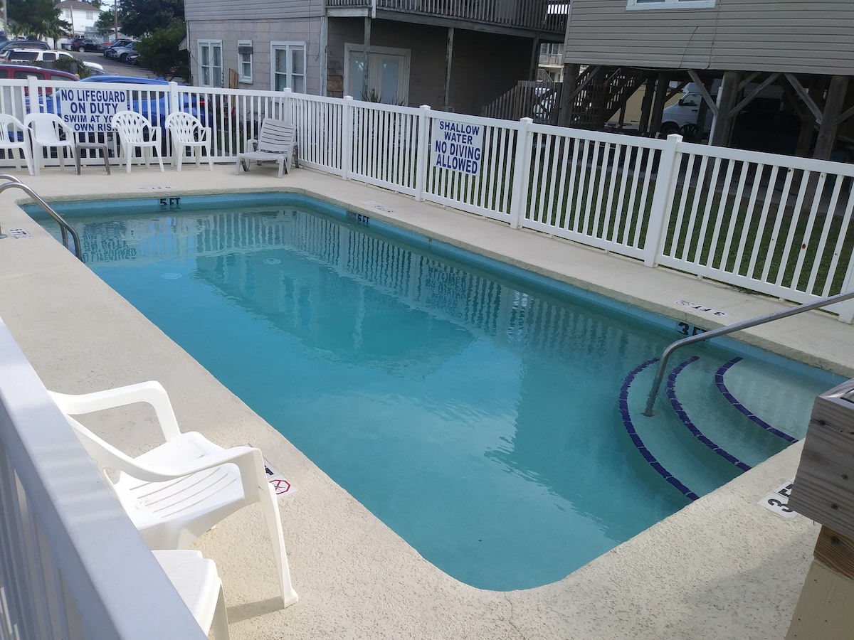 A pool area is depicted, featuring a rectangular swimming pool enclosed by a white picket fence. Several white lounge chairs are positioned along the edge, providing seating for relaxation. Clear water is visible in the pool, highlighting its inviting appearance.