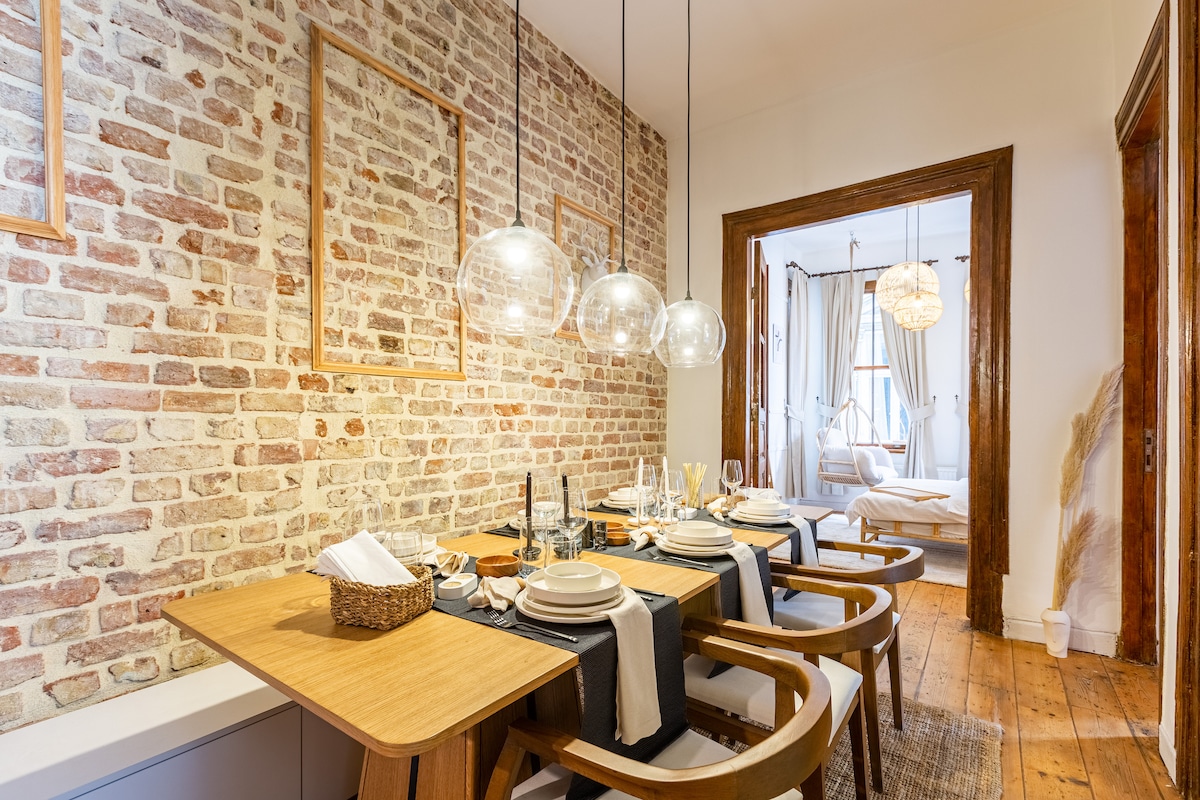 A dining area features a wooden table set for a meal, complemented by stylish chairs. Exposed brick walls provide a rustic backdrop, while modern glass pendant lights hang overhead. A glimpse of the sunlit living area is visible through the doorway at the end of the space.