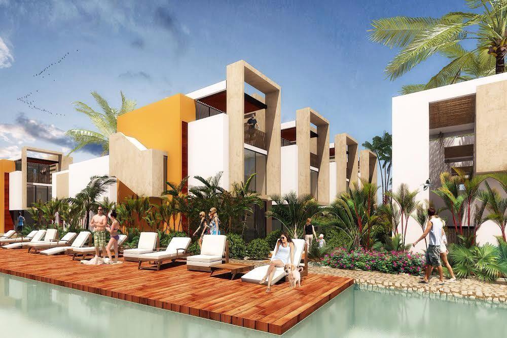 The image showcases a landscaped area with wooden decking surrounding a tranquil pool. Lounge chairs are positioned along the water's edge, and lush tropical plants create a vibrant atmosphere. The modern architecture of the villas is visible in the background, reflecting a welcoming design.
