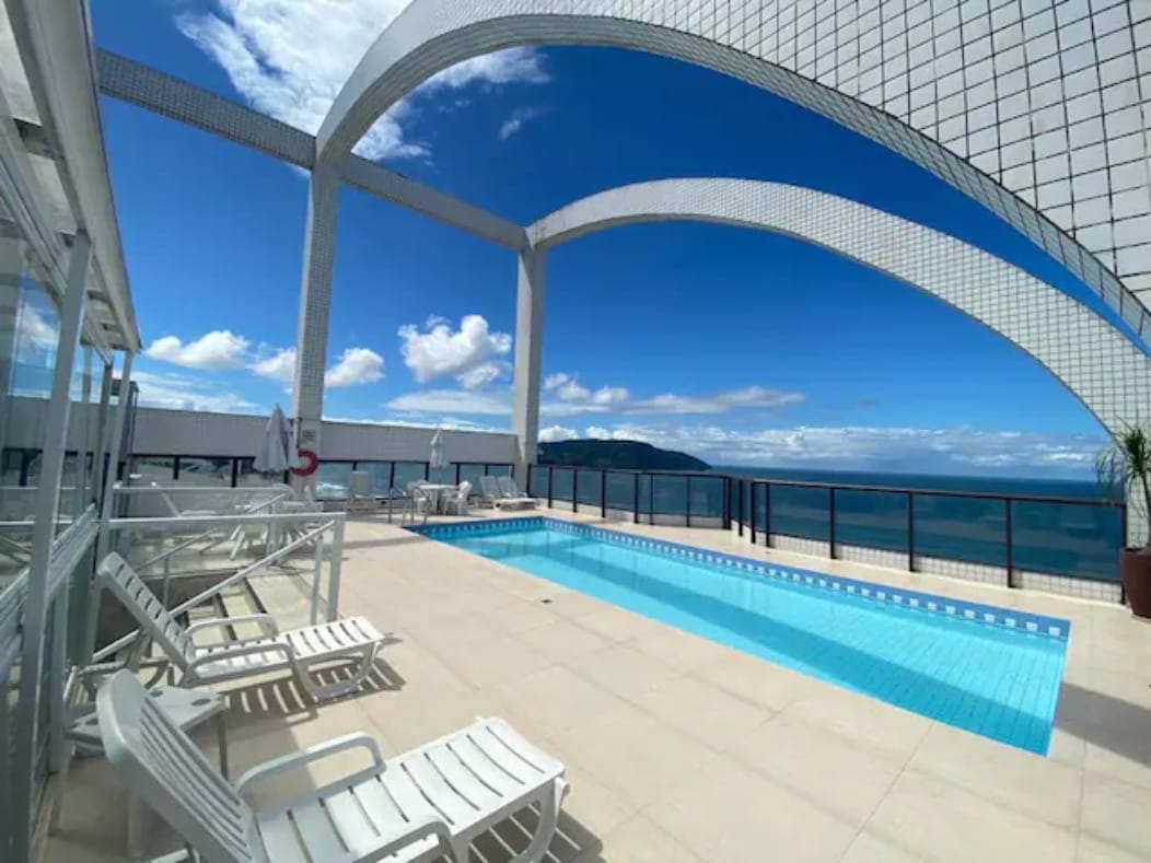 An outdoor pool area is showcased with sleek, white lounge chairs positioned beside a clear blue pool. Expansive arches frame the space, opening up to a panoramic view of the ocean and sky, creating a serene atmosphere.