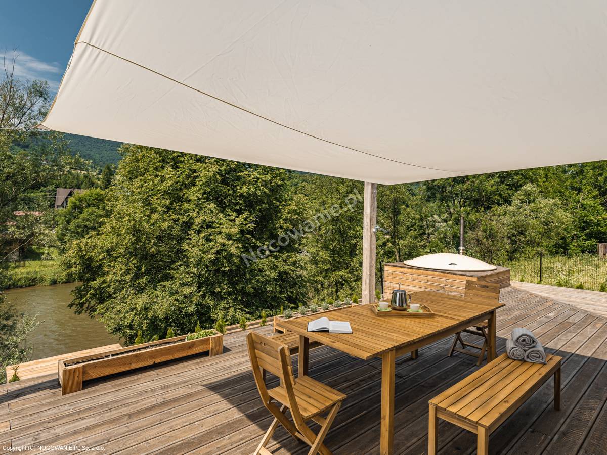 A shaded wooden terrace is presented, featuring a rectangular table surrounded by chairs. A hot tub is positioned nearby, with towels neatly arranged beside it. Lush greenery and a view of the river enhance the serene outdoor setting.