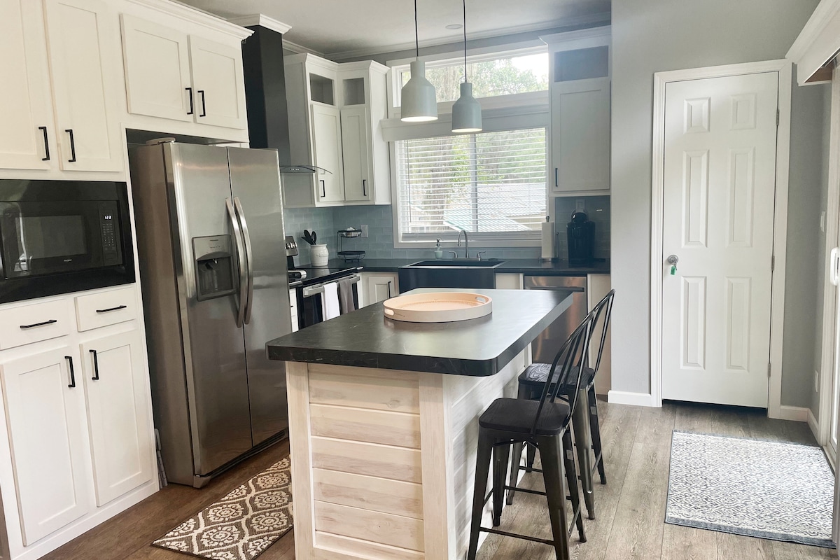 A modern kitchen features stainless steel appliances, including a refrigerator and microwave. A central island with bar stools provides additional seating. Natural light enters through a large window, illuminating the sleek cabinetry and countertops, while a patterned rug adds warmth to the space.